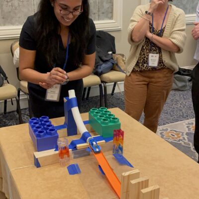 Participants eagerly watch a chain reaction contraption in action during a team building activity. A woman smiles as she sets off the colorful domino and building block setup on the table. thumbnail