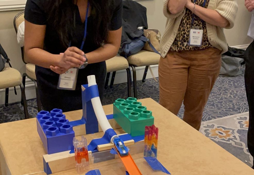 Participants eagerly watch a chain reaction contraption in action during a team building activity. A woman smiles as she sets off the colorful domino and building block setup on the table.