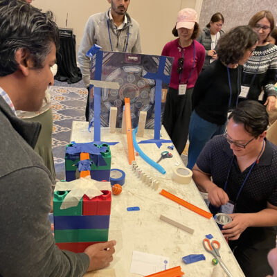 Participants collaborate to build a Rube Goldberg-inspired machine for a chain reaction team building activity, using colorful blocks, tracks, and other materials. thumbnail