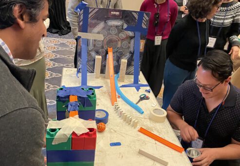 Participants collaborate to build a Rube Goldberg-inspired machine for a chain reaction team building activity, using colorful blocks, tracks, and other materials.
