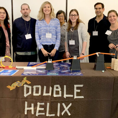 Team stands behind their completed 'Double Helix' themed chain reaction machine, showcasing their collaborative creation at a team building event. thumbnail