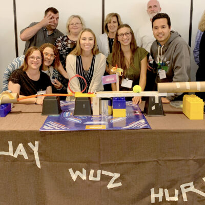 Team members gather proudly around their completed chain reaction machine at a team building event, featuring creative structures and labeled with playful text on the table. thumbnail