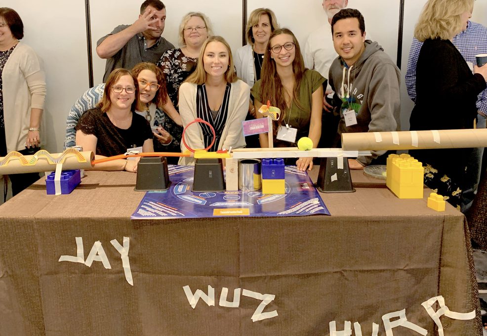 Team members gather proudly around their completed chain reaction machine at a team building event, featuring creative structures and labeled with playful text on the table.