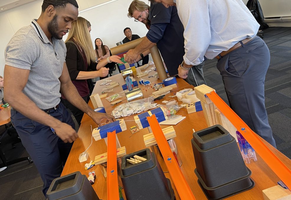 Team members collaborating during the Domino Effect team building event, working with various materials and components on a table to create complex contraptions.