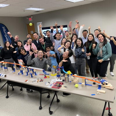 A group of excited participants celebrate their collaborative success after completing a Rube Goldberg-inspired contraption during the Domino Effect team building event. thumbnail