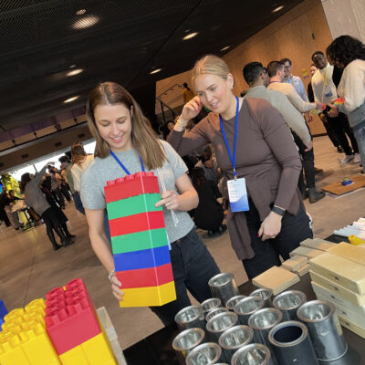 Team members work together during a chain reaction team building activity, assembling large colorful blocks as part of their Rube Goldberg-inspired machine. thumbnail