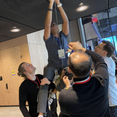 Team members work together to lift a colleague, helping him reach up to complete a part of their chain reaction machine in a collaborative team building exercise. thumbnail