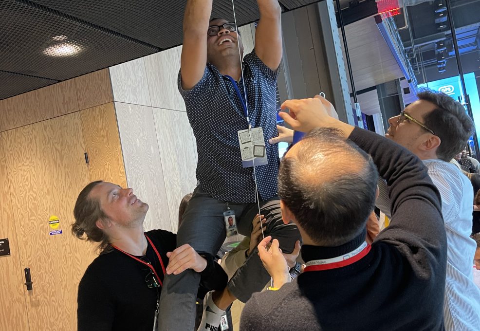 Team members work together to lift a colleague, helping him reach up to complete a part of their chain reaction machine in a collaborative team building exercise.