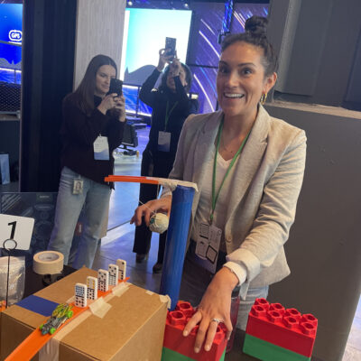Participant excitedly tests her team's chain reaction machine at a team building event, with others capturing the moment in the background. thumbnail