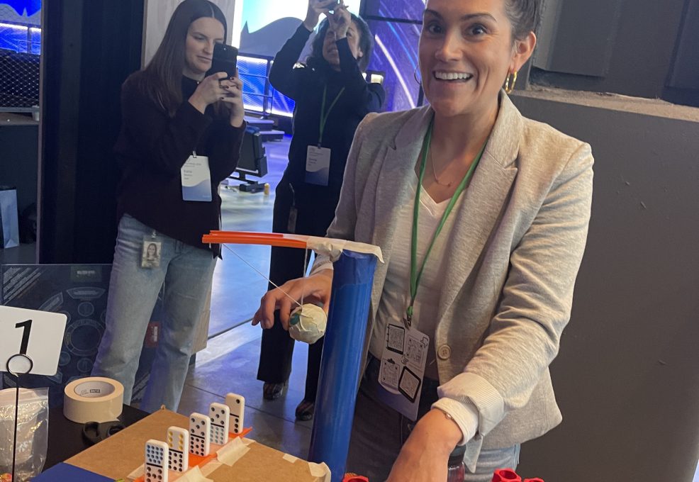 Participant excitedly tests her team's chain reaction machine at a team building event, with others capturing the moment in the background.