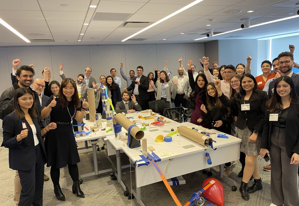 Group of participants celebrate the completion of their collaborative Chain Reaction machine during a team building event, gathered around a complex setup of interconnected parts on a table.
