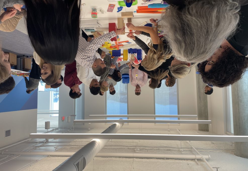 Participants in a team building activity collaborate to create a chain reaction setup. The group is fully engaged, working together with various materials on a shared table, fostering teamwork and creativity.