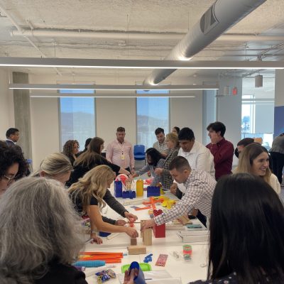 Participants in a team building activity collaborate to create a chain reaction setup. The group is fully engaged, working together with various materials on a shared table, fostering teamwork and creativity. thumbnail