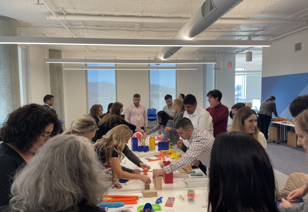 Participants in a team building activity collaborate to create a chain reaction setup. The group is fully engaged, working together with various materials on a shared table, fostering teamwork and creativity.
