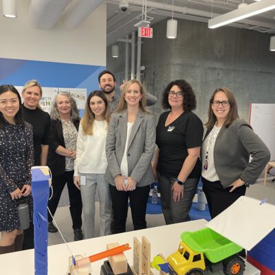 Group photo of team members standing together behind a chain reaction setup during a team building event. The participants are smiling, showcasing a collaborative and friendly atmosphere. thumbnail