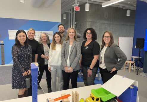 Group photo of team members standing together behind a chain reaction setup during a team building event. The participants are smiling, showcasing a collaborative and friendly atmosphere.
