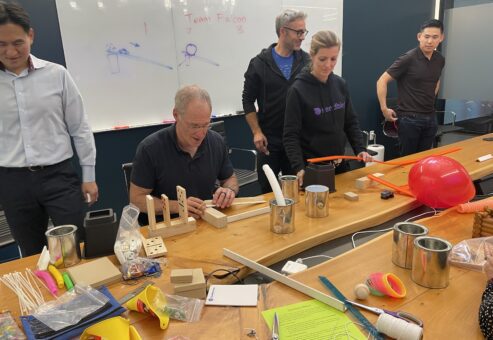 Team members collaborating and building creative contraptions during the Domino Effect team building event, surrounded by various building materials and tools.