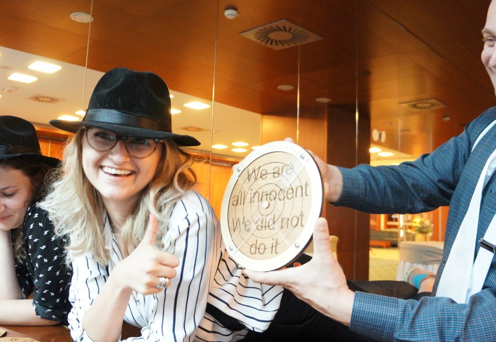 Participants in themed costumes, including hats and suspenders, holding up a clue that reads 'We are all innocent, We did not do it' during the 'Escape the Mob' team building event.