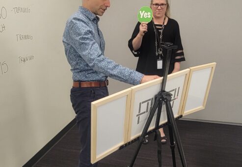 Team building exercise focused on communication and problem-solving. A participant wearing a hard hat reconstructs a model while receiving 'yes' or 'no' feedback, emphasizing the importance of clear instructions and teamwork.