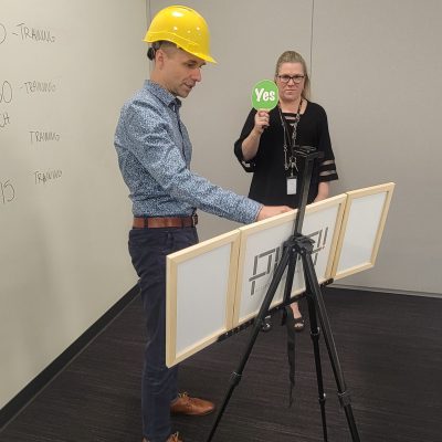 Team building exercise focused on communication and problem-solving. A participant wearing a hard hat reconstructs a model while receiving 'yes' or 'no' feedback, emphasizing the importance of clear instructions and teamwork. thumbnail