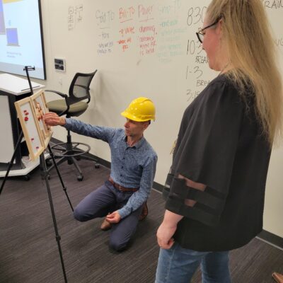 Team building activity focused on communication and problem-solving. A participant wearing a hard hat carefully assembles a model while receiving guidance, emphasizing teamwork, strategy, and clear instructions. thumbnail