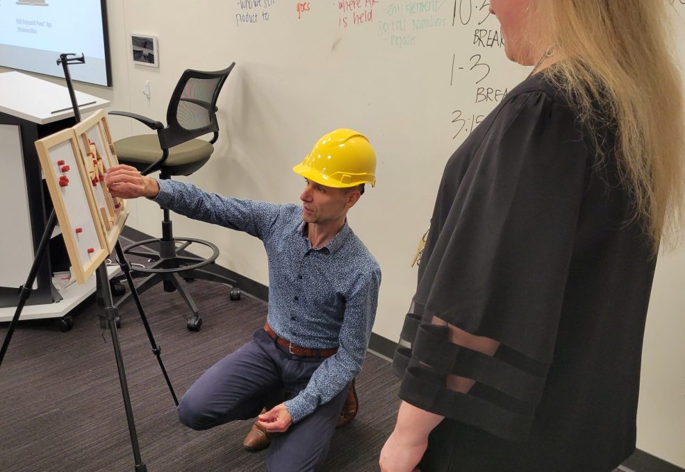 Team building activity focused on communication and problem-solving. A participant wearing a hard hat carefully assembles a model while receiving guidance, emphasizing teamwork, strategy, and clear instructions.