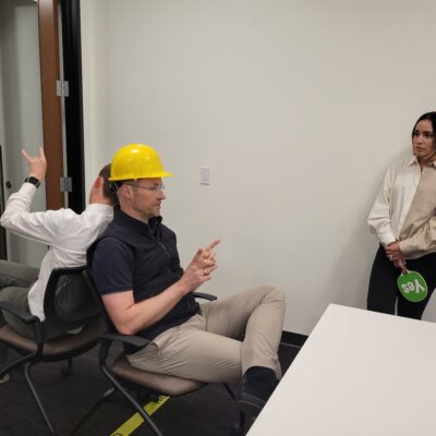 Team building exercise focused on non-verbal communication. A participant wearing a hard hat uses gestures to convey a message while a teammate, seated back-to-back, interprets the instructions, emphasizing teamwork and clear information transfer. thumbnail