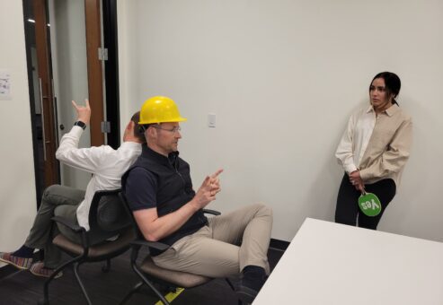 Team building exercise focused on non-verbal communication. A participant wearing a hard hat uses gestures to convey a message while a teammate, seated back-to-back, interprets the instructions, emphasizing teamwork and clear information transfer.
