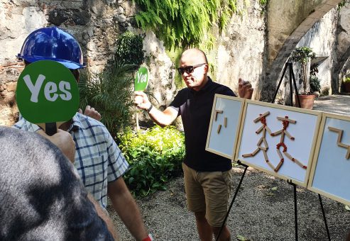 Outdoor team building activity focused on communication and accuracy. Participants use 'yes' signs to provide feedback while teammates construct a model, emphasizing teamwork, strategy, and clear instructions.