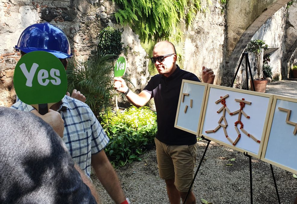 Outdoor team building activity focused on communication and accuracy. Participants use 'yes' signs to provide feedback while teammates construct a model, emphasizing teamwork, strategy, and clear instructions.