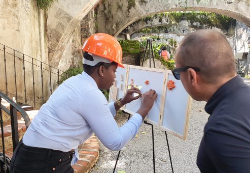 Outdoor team building exercise focused on communication and problem-solving. A participant wearing a hard hat carefully assembles a model while receiving guidance from a teammate, emphasizing teamwork and strategy.