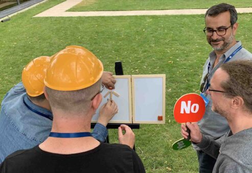 Outdoor team building activity focused on communication and feedback. Participants wearing hard hats work together to assemble a model while receiving 'yes' or 'no' responses, reinforcing teamwork and clear instruction skills.