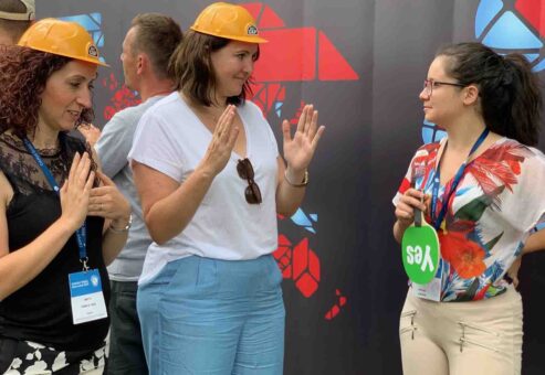 Team building activity focused on non-verbal communication. Participants wearing hard hats use gestures to convey a message while receiving feedback, emphasizing teamwork, clarity, and collaboration.