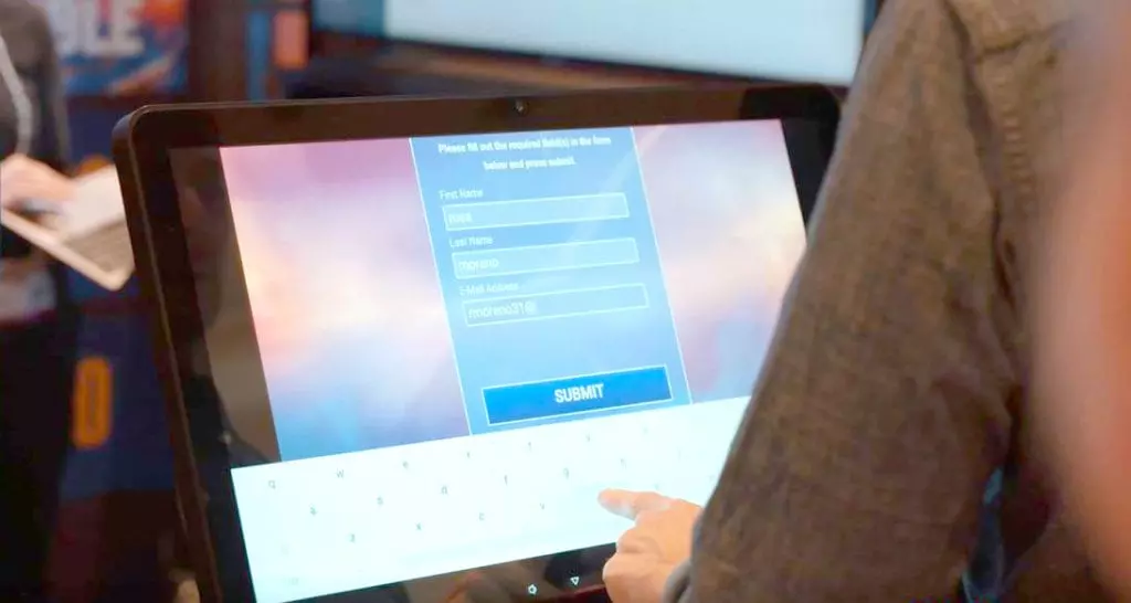 Close-up of a person filling out a form on a touchscreen tablet, entering details like 'First Name: Ross' and 'Last Name: Moreno.' A keyboard is visible on the screen, with the person typing to complete the form to start participating on the team building program: Jeopardy!