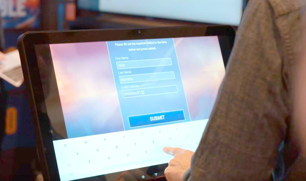 Close-up of a person filling out a form on a touchscreen tablet, entering details like 'First Name: Ross' and 'Last Name: Moreno.' A keyboard is visible on the screen, with the person typing to complete the form to start participating on the team building program: Jeopardy!