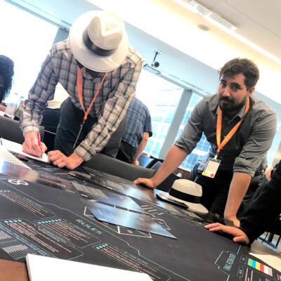 Team members collaborate on solving puzzles during The Infinite Loop team building event, with one participant writing notes and another observing the challenge map. thumbnail