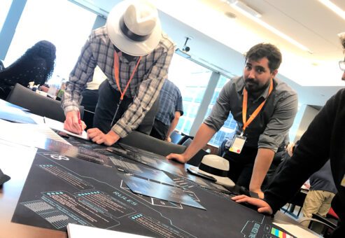 Team members collaborate on solving puzzles during The Infinite Loop team building event, with one participant writing notes and another observing the challenge map.
