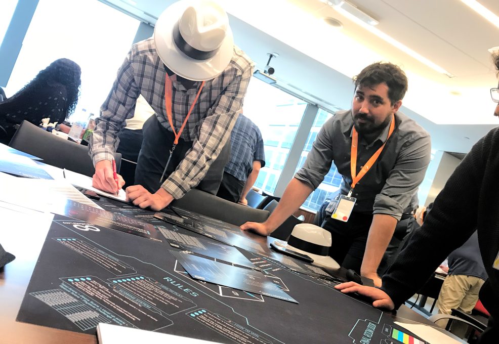 Team members collaborate on solving puzzles during The Infinite Loop team building event, with one participant writing notes and another observing the challenge map.
