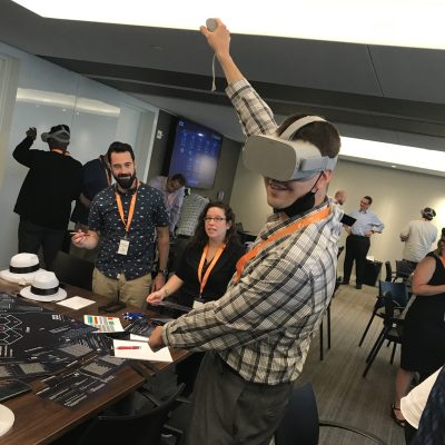 A participant wearing a VR headset raises his arm while engaging in The Infinite Loop team building activity, surrounded by teammates and facilitators in a collaborative environment. thumbnail