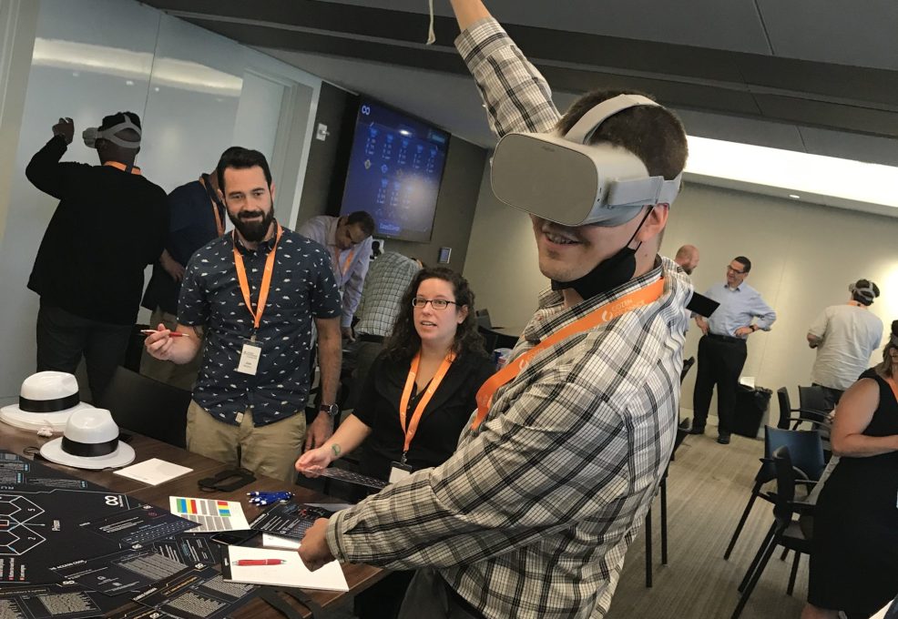 A participant wearing a VR headset raises his arm while engaging in The Infinite Loop team building activity, surrounded by teammates and facilitators in a collaborative environment.