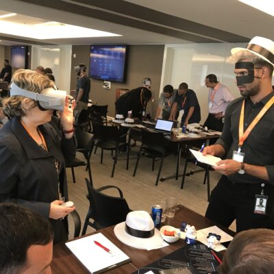 Participants at The Infinite Loop team building event engage in problem-solving activities, with one person wearing a VR headset and another taking notes while wearing a themed mask and hat. thumbnail