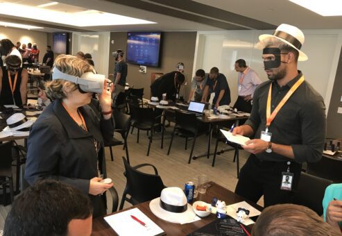 Participants at The Infinite Loop team building event engage in problem-solving activities, with one person wearing a VR headset and another taking notes while wearing a themed mask and hat.
