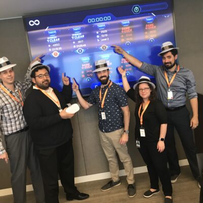 A team celebrates their progress, pointing at a virtual leaderboard during The Infinite Loop team building event, while wearing themed hats and badges. thumbnail