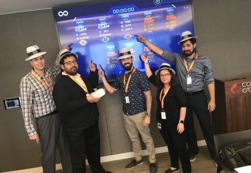 A team celebrates their progress, pointing at a virtual leaderboard during The Infinite Loop team building event, while wearing themed hats and badges.