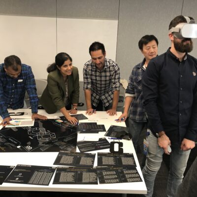 Team members collaborate at a table while one participant engages with a virtual reality headset during The Infinite Loop team building event. thumbnail