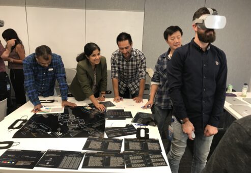 Team members collaborate at a table while one participant engages with a virtual reality headset during The Infinite Loop team building event.