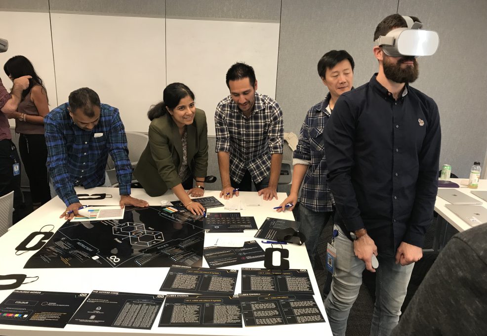 Team members collaborate at a table while one participant engages with a virtual reality headset during The Infinite Loop team building event.