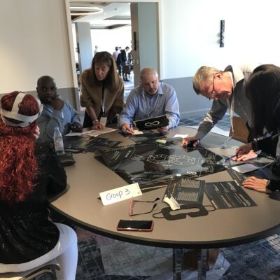 A group of participants collaborating on puzzle tasks at The Infinite Loop team building event, while one team member wears a VR headset to solve virtual challenges. thumbnail