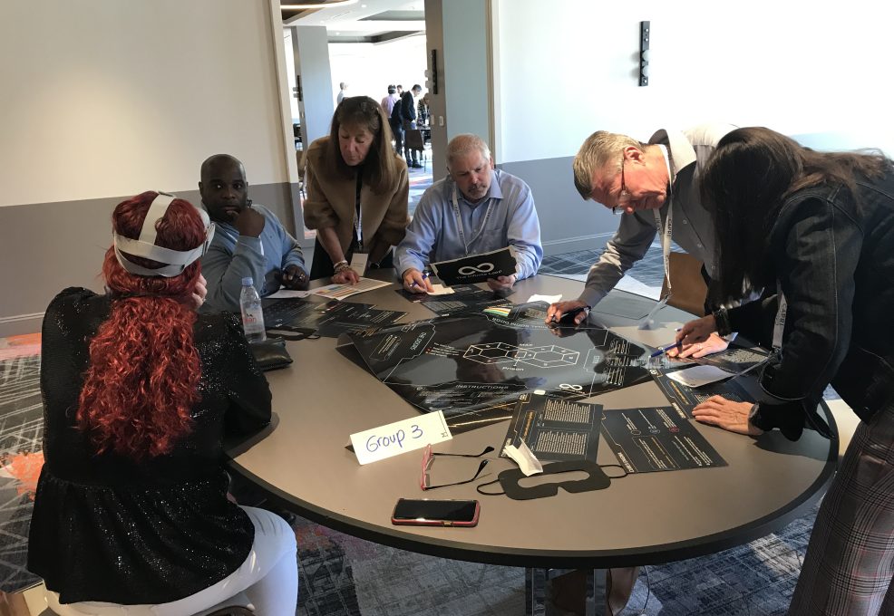 A group of participants collaborating on puzzle tasks at The Infinite Loop team building event, while one team member wears a VR headset to solve virtual challenges.