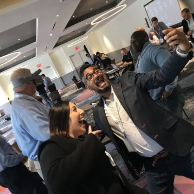 Two participants enthusiastically taking a selfie during The Infinite Loop team building event, while others engage in VR challenges in the background. thumbnail